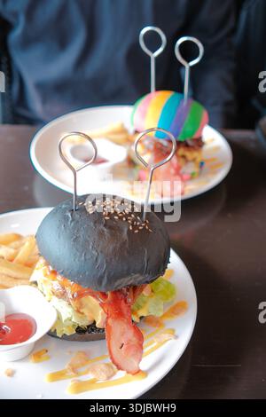 Handmade gourmet hamburger with black bun, bacon, cheese, and fresh vegetables served with french fries on a plate. Stock Photo