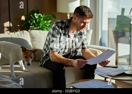 Man sitting on couch at home examining paper bills and financial documents near a laptop while dog standing nearby. Concept of managing personal finan Stock Photo