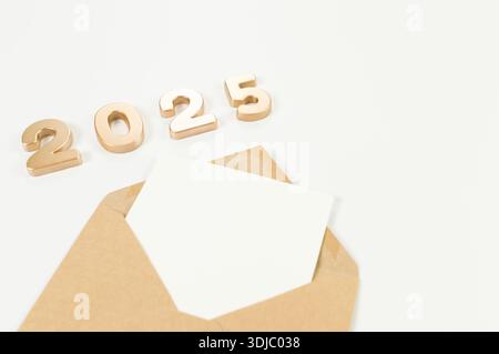 2025 Happy New Year. Top view of gold numbers, brown kraft envelope, white card on white background. Copy space, flat lay. Stock Photo