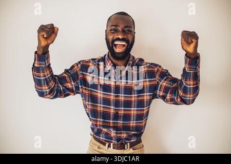 Cheerful black man in cozy shirt shirt standing in front of wall and smiling. He is raising his arms in euphoria. Stock Photo