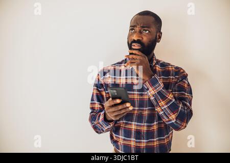 Anxious black man in cozy shirt standing in front of wall and typing messages smartphone. Stock Photo