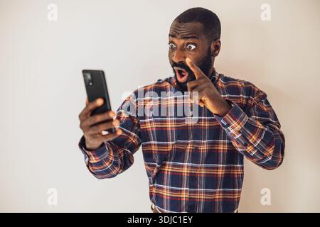 Surprised and shocked black man in cozy shirt standing in front of wall and having video call on smartphone. Stock Photo
