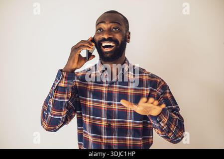 Cheerful black man in cozy shirt standing in front of wall and talking on mobile phone. Stock Photo