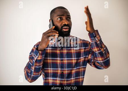 Anxious black man in cozy shirt standing in front of wall and talking on mobile phone. Stock Photo