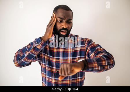 Anxious black man in cozy shirt standing in front of wall and looking at his wristwatch. He will be late. Stock Photo