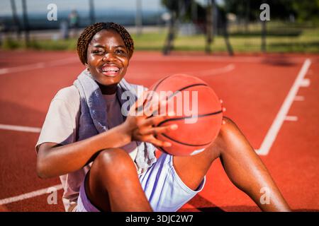 Young black woman is sitting on basketball court. She is holding the ball. Stock Photo