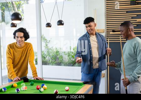 Diverse male friends leaning, holding cues, preparing shot around green pool table in rec room Stock Photo