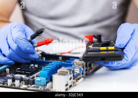 Technician repairs computer motherboard with multimeter and tools in workshop setting. Stock Photo
