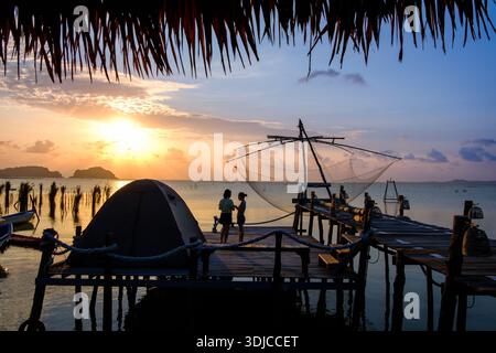Overhead rear view of a woman sitting on a wooden jetty with two kayaks moored alongside at sunset, Ko Mat, Songkhla Lake, Phatthalung, Thailand Stock Photo