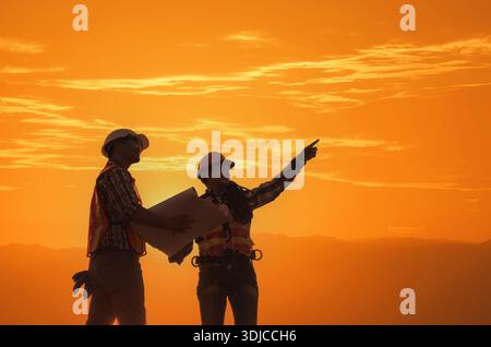 Silhouette of Two construction workers looking at plans on a construction site at sunset, Thailand Stock Photo