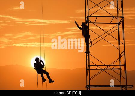 Silhouette of a construction worker hanging in a harness with another person standing on structural steelwork at sunset, Thailand Stock Photo