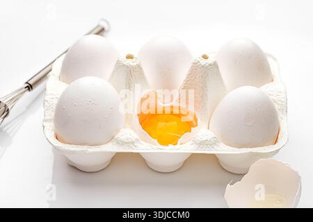 Close-up of a carton of six eggs with one cracked open on a table with a metal whisk Stock Photo