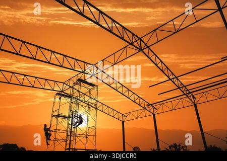 Silhouette of two construction workers climbing on structural steelwork at sunset, Thailand Stock Photo