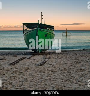 A vibrant green fishing boat rests on a sandy beach, hauled ashore with a rope and wooden rails. The tranquil scene is bathed in the warm glow of a su Stock Photo