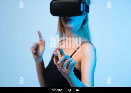 Woman wearing VR headset and interacting with virtual interface using hand gestures in modern studio lighting Stock Photo