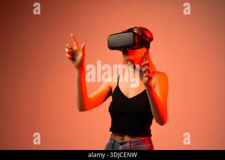 Young woman using virtual reality headset and interacting with digital interface using hand gestures in warm studio lighting Stock Photo