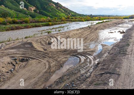 Murky river flows through a wide, sandy riverbed with numerous vehicle tracks and reflective puddles, backed by verdant hills under a cloudy sky. Stock Photo