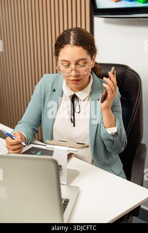 Professional woman at desk multitasking, writing on a document and holding a mobile phone Stock Photo