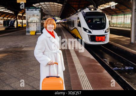 Caucasian redhead woman with orange suitcase standing on railway platform Stock Photo