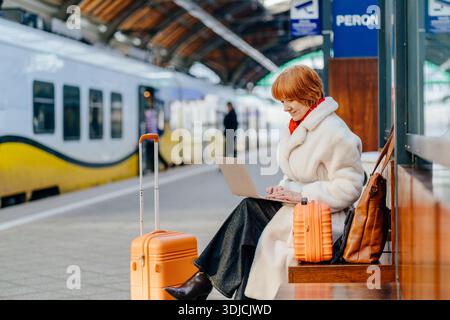 Successful middle aged caucasian redhead woman working on laptop at train station platform Stock Photo