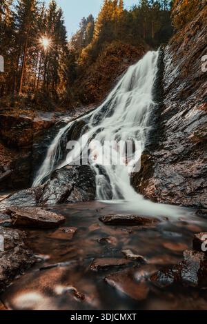 Long exposure view of the Valul Miresei waterfall cascading over rocky terrain through a pine forest with a sunstar shining through the trees in the A Stock Photo