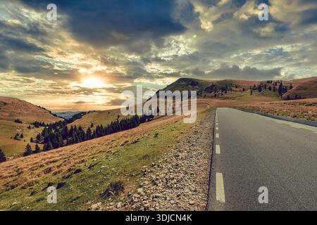 Wide angle view of the empty Transbucegi mountain road winding through the rolling grassy hills under a dramatic cloudy sunset sky in the Bucegi Mount Stock Photo