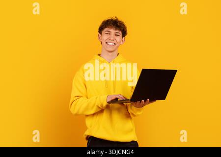 Cheerful young man using laptop on bright yellow background, representing remote work Stock Photo