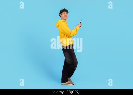 Smiling young man in yellow hoodie holding smartphone with surprised expression Stock Photo