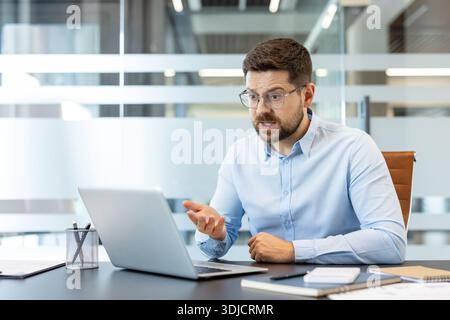Businessman looking frustrated and stressed, encountering a problem while working remotely on his laptop from the office desk, expressing confusion and trouble online Stock Photo