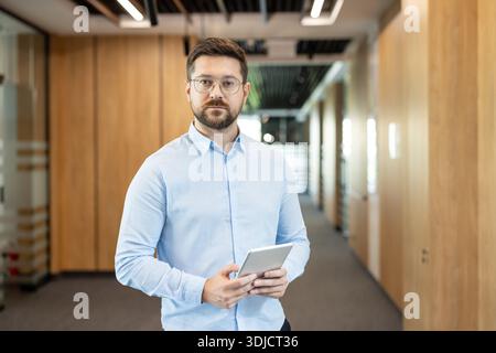 Young businessman wearing a blue shirt and eyeglasses standing confidently in a modern office corridor, holding a digital tablet and looking directly at the camera Stock Photo