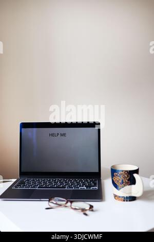 Laptop on white desk with message Help me on screen, coffee cup and glasses. Concept of online support, technical problem and remote work stress. Stock Photo