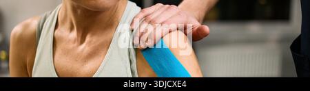 Header of Middle aged Caucasian woman receiving physical therapy, therapist applying blue kinesiology tape to shoulder, close up of therapist hand on patient shoulder during rehabilitation session Stock Photo
