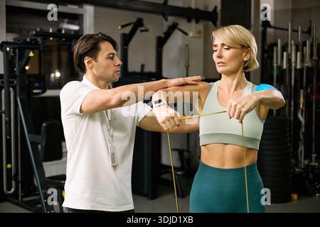 Caucasian young adult woman performing resistance band shoulder exercise under supervision of Caucasian young adult man physical therapist in rehabilitation center gym setting Stock Photo