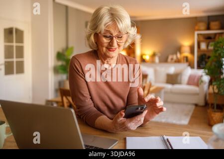Senior woman with laptop on desk using mobile phone to pay household bills online while sitting at home Stock Photo