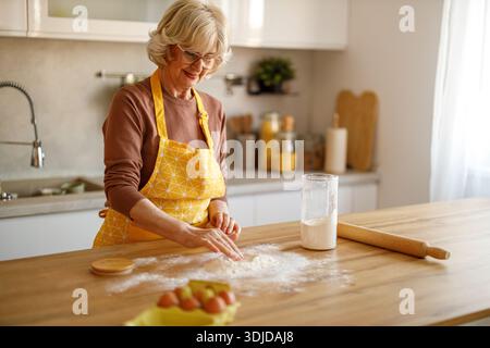 Happy senior woman in eyeglasses kneading dough and preparing homemade sweets on kitchen island at home Stock Photo