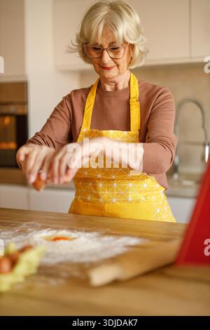 Senior woman in apron and eyeglasses cracking eggs into flour on island while sharing recipe on blog Stock Photo