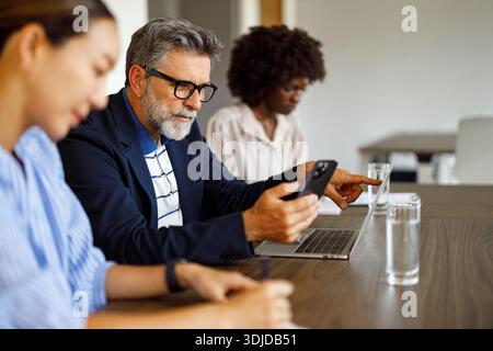 Male professional texting on phone and using laptop while collaborating with female colleagues in modern office Stock Photo