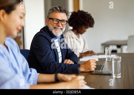 Confident male lawyer smiling at camera while working on laptop and sitting with female professionals in modern office Stock Photo