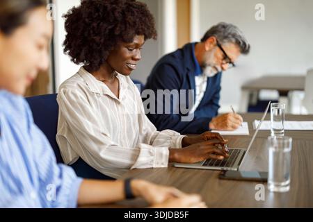 Smiling businesswoman with afro hair working on laptop while colleagues analyzing documents together at desk in office Stock Photo