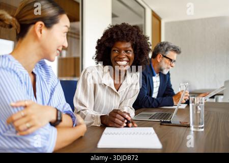 Smiling female lawyers collaborating documents while businessman reviews paperwork in background during office meeting Stock Photo