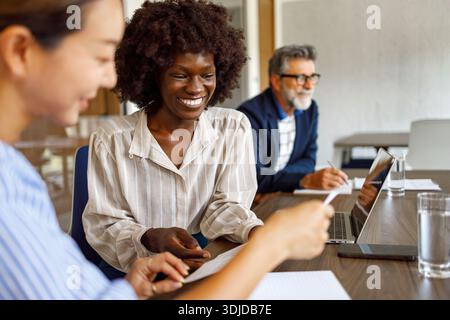 Smiling female professionals reviewing financial documents while businessman collaborates in background in modern office Stock Photo