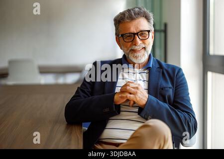 Portrait of bearded male professional in eyeglasses smiling at camera while sitting confidently in boardroom Stock Photo