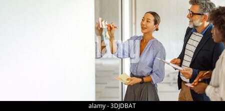 Businesswoman writing ideas on sticky note while collaborating with teammates during creative meeting Stock Photo