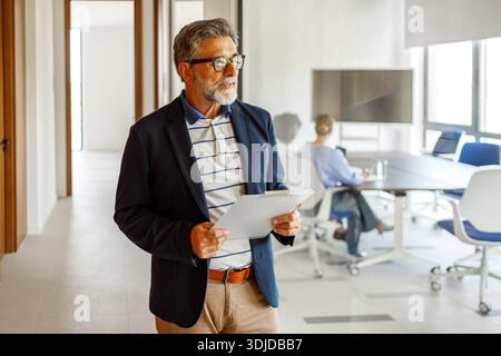 Thoughtful mature businessman in eyeglasses holding documents and looking away while working in modern office Stock Photo