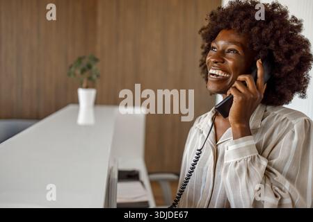 Smiling female receptionist talking on phone with guest and making reservation at hotel reception desk Stock Photo