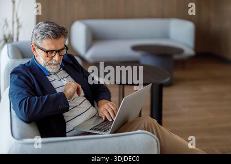 Focused bearded male professional doing online research over laptop while sitting on sofa in corporate office Stock Photo