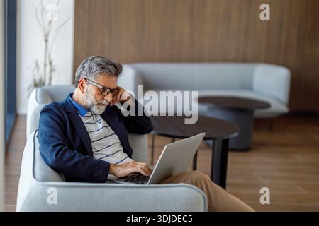 Focused businessman in eyeglasses sitting on sofa in modern office working on laptop analyzing financial data Stock Photo