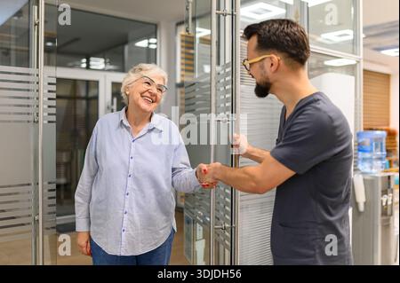 Senior woman offering handshake to cheerful male doctor opening door welcoming her into hospital for medical appointment Stock Photo