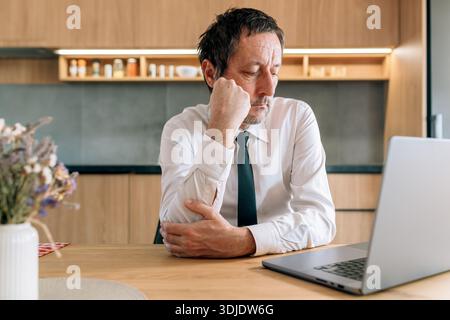 Portrait of a businessman sitting at a modern kitchen home office desk, head bowed with worried expression, selective focus Stock Photo