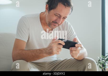 Adult male enjoying mobile gaming on smartphone in warm morning home interior, relaxed casual mood and cozy atmosphere. Selective focus. Stock Photo